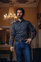 Man wearing a patterned shirt and jeans standing in a room with a chandelier.
