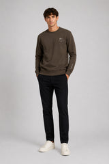 A man modeling a dark brown crewneck sweatshirt with a small white chest graphic, paired with black trousers and white sneakers, against a light gray studio background.