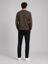 A man viewed from behind wearing a dark brown crewneck sweatshirt, black trousers, and white sneakers, standing against a neutral light gray studio background.