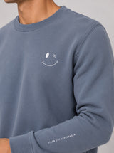 A close-up of a person wearing a dusty slate blue crewneck sweatshirt with a white minimalist smiley face on the chest and 'CLEAN CUT COPENHAGEN' printed on the left sleeve.