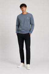 A man modeling a slate blue crewneck sweatshirt with a small white embroidered smiley face logo on the left chest, paired with black trousers and white sneakers, against a light gray studio background.