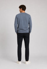 A man viewed from behind wearing a dusty slate blue crewneck sweatshirt, black trousers, and white sneakers, standing against a light gray studio background.