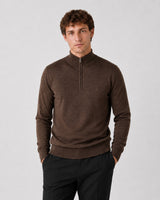 A man wearing a dark brown quarter-zip knit sweater and black trousers, standing against a light gray studio background.