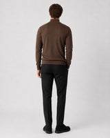 A man viewed from behind wearing a brown mock-neck knit sweater and black trousers, standing against a light gray studio background.