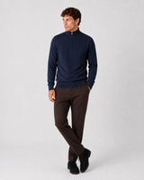 A man modeling a navy blue quarter-zip pullover sweater and dark brown trousers against a light gray studio background.