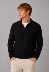 A man wearing a black long-sleeve polo sweater over a white t-shirt and beige trousers, standing against a neutral gray studio background.