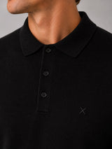 Close-up of a man wearing a black ribbed knit polo shirt with a three-button placket and a small tonal embroidered logo on the chest.