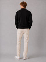 A man is shown from behind wearing a black long-sleeve knit polo shirt, off-white chino pants, and white sneakers with red stripes, standing against a neutral gray studio background.