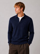 A man wearing a navy blue long-sleeve polo sweater over a white t-shirt and brown trousers, standing against a neutral gray studio background.