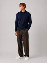 A man wearing a navy blue long-sleeve polo sweater, dark brown trousers, and white sneakers, standing against a neutral light gray studio background.