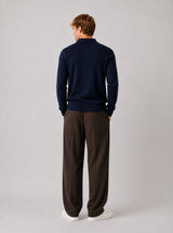 A man viewed from behind wearing a navy blue mock-neck knit sweater and dark brown wide-leg trousers, standing against a neutral light gray studio background.