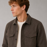 A man wearing a dark brown textured overshirt over a cream ribbed knit shirt, shown in a close-up profile shot against a neutral gray background.