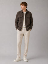 A man wearing a dark brown overshirt, cream-colored trousers, a cream crewneck shirt, and white sneakers, standing against a solid gray studio background.