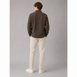 A man is shown from behind wearing a dark brown long-sleeve button-up shirt, off-white trousers, and white sneakers, standing against a solid gray studio background.