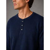 A close-up of a man wearing a navy blue waffle-knit henley shirt with three buttons on the chest, set against a neutral gray background.