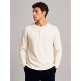 A man wearing a cream-colored, long-sleeve ribbed henley shirt and dark pants, standing against a neutral gray studio background.
