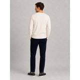 A man is shown from behind wearing a cream long-sleeve crewneck sweater, dark navy trousers, and navy loafers, standing against a neutral gray studio background.