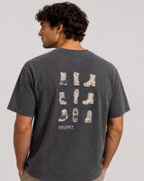 A man wearing a dark gray washed t-shirt, viewed from the back. The shirt features a graphic print of various boot silhouettes in a light beige color on the back, with the brand name 'EXPLORE+' printed below.