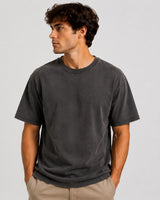 A man wearing a washed black short-sleeve crewneck t-shirt and khaki pants, standing against a light gray studio background.