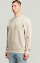 Man wearing a beige sweatshirt and light blue jeans against a plain background