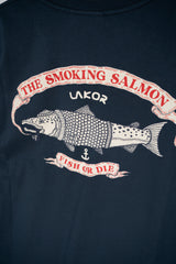 Close-up of a dark navy blue t-shirt featuring a graphic print of a salmon with a smoking pipe, framed by banners reading 'THE SMOKING SALMON' and 'FISH OR DIE', with 'LAKOR' printed above.