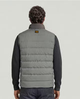 A man viewed from behind wearing a gray quilted puffer vest over a black long-sleeve shirt, against a light gray studio background.