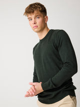 A young man with light brown hair wearing a dark forest green long-sleeve crewneck sweater and beige trousers, posing against a light gray studio background.