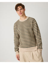 A man wearing a beige and dark green geometric patterned knit sweater with beige trousers, photographed against a light gray studio background.