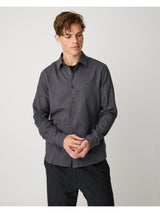 A man wearing a dark gray long-sleeve button-up shirt and dark trousers, standing against a light gray studio background.