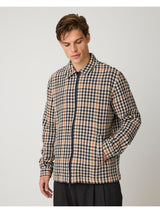 A man wearing a long-sleeve zip-up jacket with a navy, beige, and brown houndstooth pattern, paired with black trousers, against a plain light gray background.