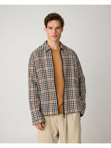 A man wearing a navy, beige, and brown houndstooth patterned overshirt over a tan ribbed sweater and beige trousers, standing against a light gray studio background.