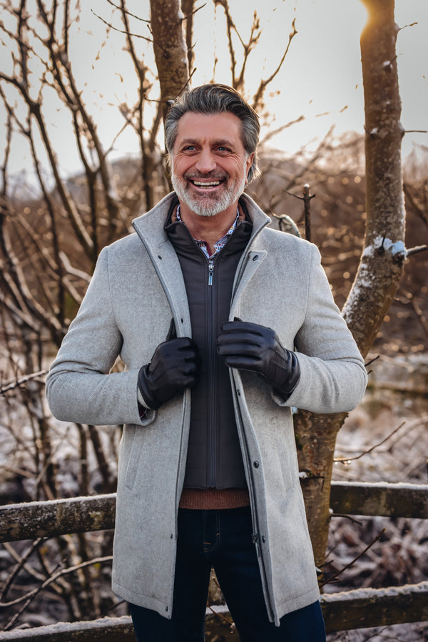 Man wearing a gray coat standing in a snowy landscape with bare trees.