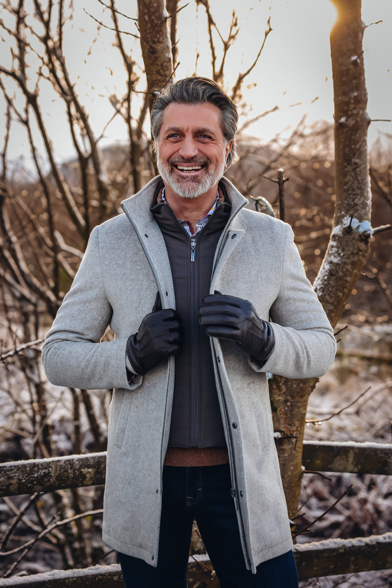 Man wearing a gray coat standing in a snowy landscape with bare trees.