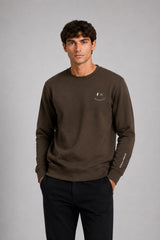 A man wearing a dark brown crewneck sweatshirt with a small white embroidered smiley face logo on the left chest, paired with black pants, standing against a solid gray studio background.