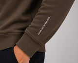 Close-up of a person wearing an olive brown sweatshirt, showing the left sleeve with white vertical text reading 'CLEAN CUT COPENHAGEN' and a ribbed cuff, paired with dark pants against a light gray background.