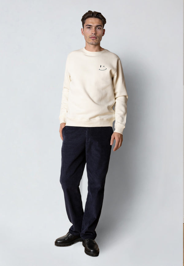 Man wearing a cream sweatshirt with a small graphic, dark pants, and black shoes on a light gray background