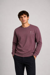 A man wearing a dusty mauve crewneck sweatshirt with a small white embroidered logo on the left chest, paired with dark pants, standing against a neutral gray studio background.