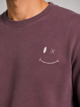 A close-up of a person wearing a crewneck sweatshirt in a muted plum-brown color, featuring a small white embroidered smiley face with X eyes on the left chest.