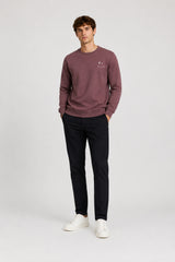 A man modeling a crewneck sweatshirt in a dusty rose color, paired with dark trousers and white sneakers, standing against a light gray studio background.