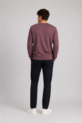 A man viewed from behind wearing a dusty mauve crewneck sweater, dark navy trousers, and white sneakers, standing against a light gray studio background.