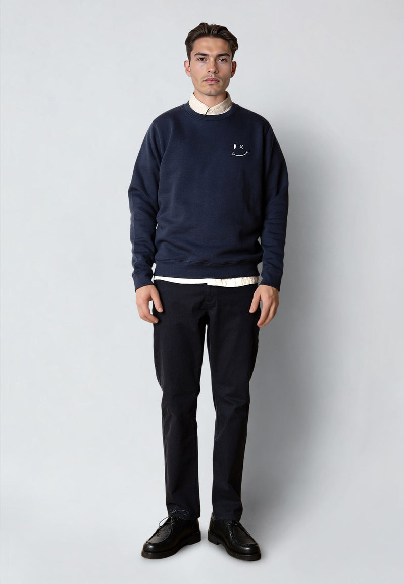 Man wearing a navy sweater with a smiley face logo, white shirt, black pants, and black shoes on a light gray background