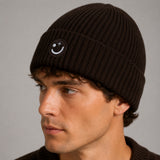 A man wearing a dark brown ribbed knit beanie with a small embroidered smiley face logo on the front, photographed against a neutral gray background.