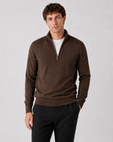 A man wearing a brown quarter-zip knit sweater over a white t-shirt and black trousers, standing against a light gray studio background.