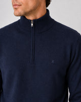 A close-up of a man wearing a navy blue quarter-zip knit sweater with a small embroidered logo on the chest, set against a plain light gray background.