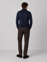 A man viewed from behind wearing a navy blue mock-neck sweater and dark brown trousers, standing against a light gray studio background.