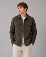 A man wearing a dark brown textured button-up overshirt jacket over a cream t-shirt and beige trousers, standing against a solid gray studio background.