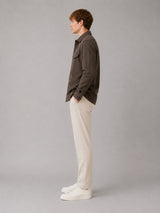A man in profile wearing a dark brown corduroy shirt jacket, off-white trousers, and white sneakers, standing against a solid gray studio background.