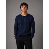 A man wearing a navy blue long-sleeve henley shirt with a three-button placket and dark trousers, standing against a solid gray studio background.