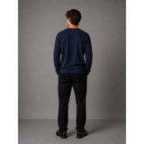 A man viewed from behind wearing a navy blue long-sleeve crewneck sweater, black trousers, and black shoes, standing against a neutral gray studio background.