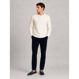 A man wearing a cream long-sleeve henley shirt, dark navy trousers, and navy boat shoes, standing against a neutral gray studio background.